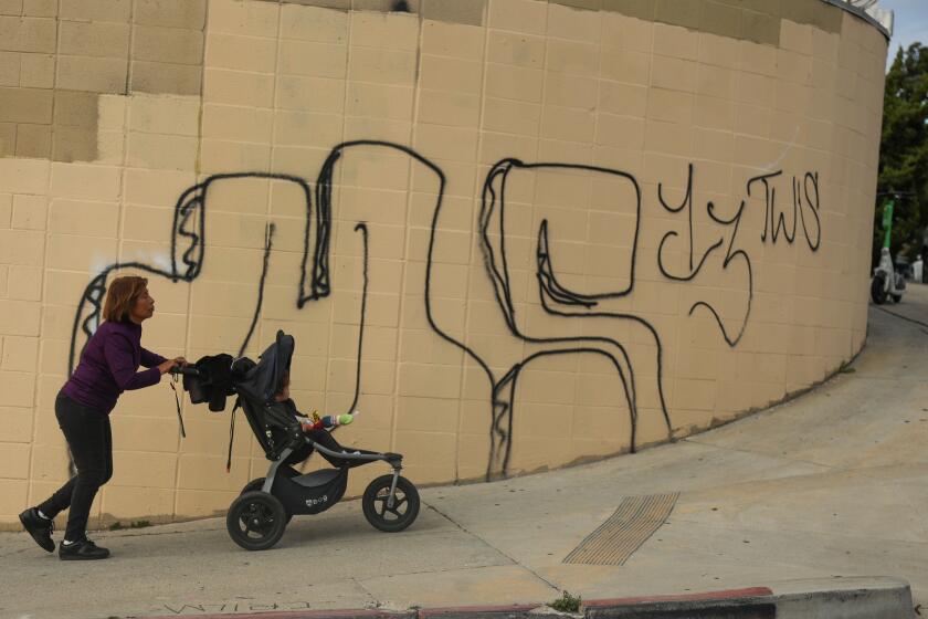 East Hollywood, CA - March 29: A woman pushes a baby stroller with a young baby near a MS-13 graffiti on Friday, March 29, 2024 in East Hollywood, CA. (Michael Blackshire / Los Angeles Times)