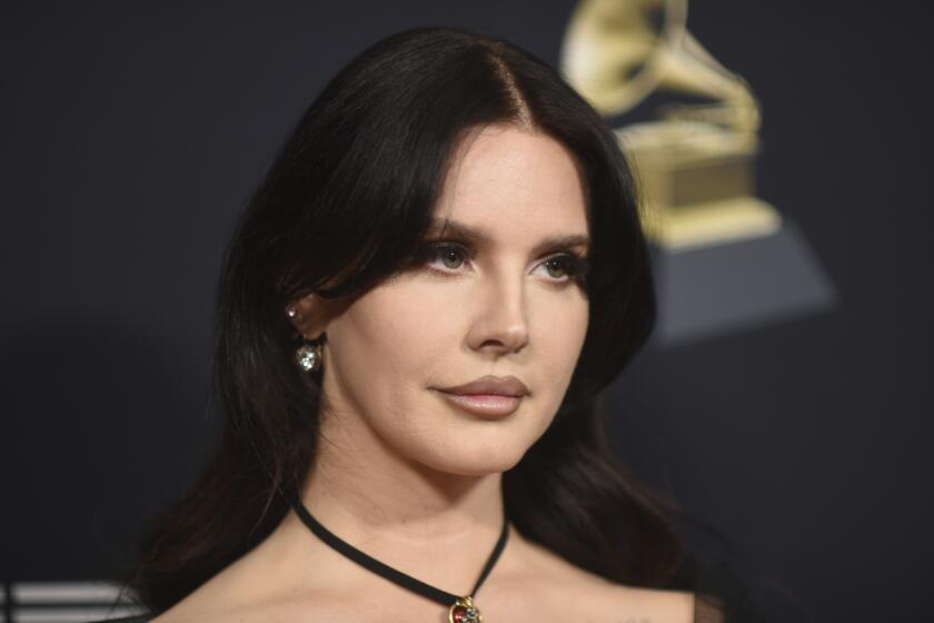 Lana Del Rey with dark hair posing against a dark background decorated with a Grammy award